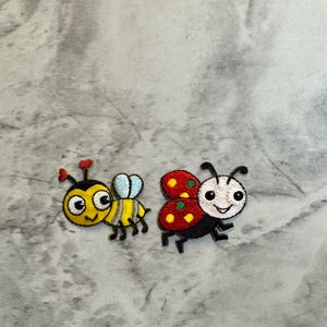 May include: Two embroidered patches: a yellow and black bee with blue wings and a red, black, and white ladybug. Both patches have smiling faces and are on a gray and white marble background.