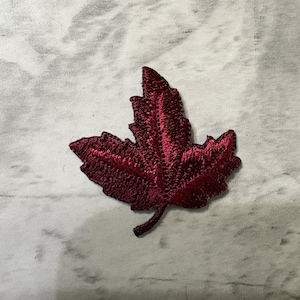 May include: A deep burgundy embroidered patch in the shape of a maple leaf. The patch features intricate thread work that creates a textured surface, highlighting the veins and jagged edges of the leaf, complete with a small stem at the base.