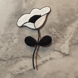 May include: Embroidered flower patch featuring a white petal design with a black center and stem. The patch includes two black leaves at the base of the stem. The flower is a decorative element.