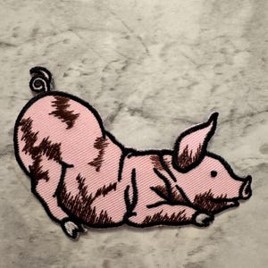 May include: A pink embroidered patch of a pig in a relaxed pose. The pig has brown markings and a black outline. The pig's curly tail is visible. The patch is designed to be ironed on or sewn onto fabric.