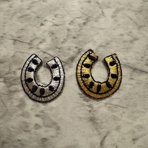 May include: Two embroidered horseshoe patches. One is silver with black accents, the other is gold with black accents. Both have a black border and are on a marble-patterned surface. These patches could be used for clothing or craft projects.