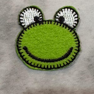 May include: A green frog-shaped patch with a smiling mouth and large, white eyes. The frog's body is a bright green, with black stitching outlining the shape and features. The eyes have black pupils and are outlined with black stitching.