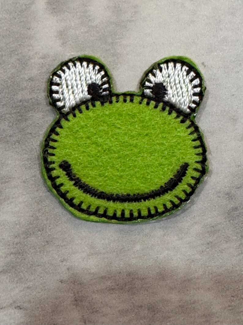 May include: A green frog-shaped patch with a smiling mouth and large, white eyes. The frog's body is a bright green, with black stitching outlining the shape and features. The eyes have black pupils and are outlined with black stitching.