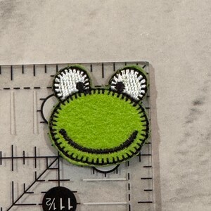 May include: A green felt frog applique with black stitching and white and black eyes. The frog is approximately 1.5 inches wide and is on a clear ruler.
