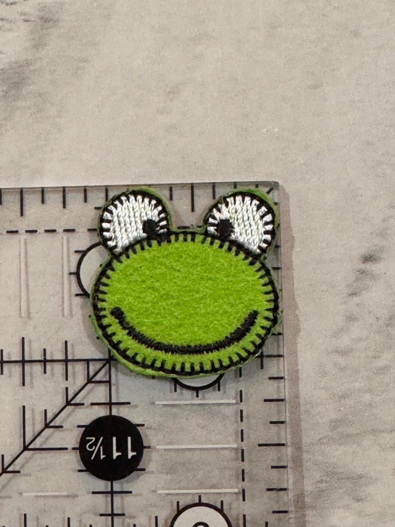 May include: A green felt frog applique with black stitching and white and black eyes. The frog is approximately 1.5 inches wide and is on a clear ruler.