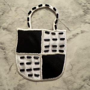 May include: A white and black patch in the shape of a shield with a handle. The shield is divided into four sections, two black and two white, with black beads sewn on the white sections and around the handle.