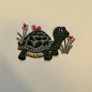 May include: A green embroidered turtle with a pink bow on its shell, surrounded by green grass and pink flowers.