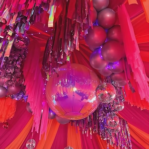 May include: A vibrant party scene with a pink and orange color scheme. The ceiling is decorated with pink streamers, balloons, and disco balls. The overall effect is festive and celebratory.