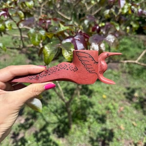 May include: A hand-carved wooden smoking pipe in a reddish-brown hue, featuring a stylized bird design on the bowl and intricate geometric patterns etched along the stem. The pipe has a smooth finish and a distinct, elongated shape.