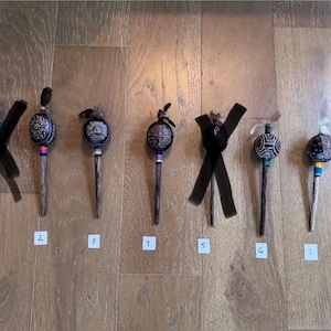 Può includere: Sette bastoncini decorativi per capelli con top sferici ornati di colore scuro e lunghi steli di legno sono esposti su una superficie di legno. I top presentano disegni intricati e sono accentuati da piume o nastri. Ogni bastoncino ha un design unico.