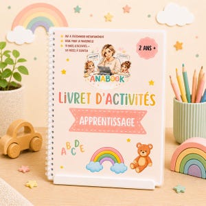 Può includere: Un libro di attività rilegato a spirale con il titolo "LIVRET D'ACTIVITÉS APPRENTISSAGE" e il testo "2 ANS +". La copertina presenta illustrazioni di una donna con bambini, un arcobaleno, un orsacchiotto e stelle. Il libro è appoggiato su un supporto bianco.