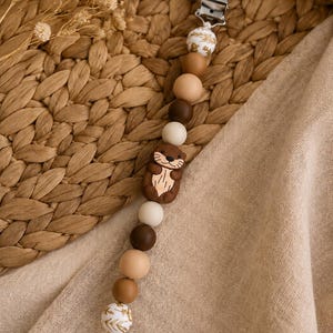 May include: A dummy clip featuring a series of round beads in shades of brown, tan, and cream, accented by two white beads with gold leaf patterns. A central brown otter-shaped bead sits in the middle, attached to a white plastic clip.
