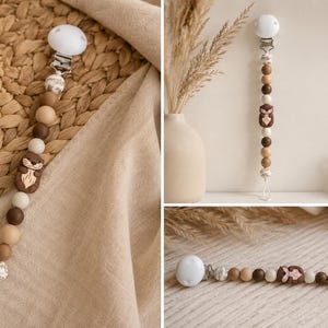 May include: A dummy clip featuring a series of round beads in shades of cream, tan, and dark brown, accented by a central brown silicone otter-shaped bead. The clip includes a white plastic fastener and a white cord loop at the bottom end.