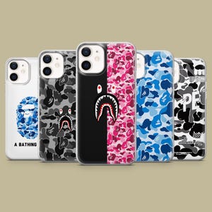 May include: A collection of phone cases with various designs. The cases feature camouflage patterns in black, gray, blue, and pink, along with a shark graphic and the text "A BATHING". The cases are designed to fit a smartphone.