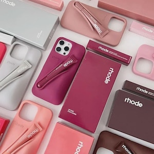 Può includere: Un assortimento di prodotti di bellezza, tra cui tinte per labbra e custodie per telefoni, in varie tonalità di rosa e marrone. I prodotti sono disposti su una superficie bianca, con il nome del marchio "rhode" visibile.