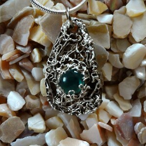 May include: A silver pendant featuring a teal green gemstone set in a wire-wrapped design. The pendant is photographed against a background of small, white, iridescent stones.