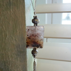 May include: A silver chain necklace with a rectangular pendant featuring a cloudy, brown, and white agate stone. The pendant is accented with silver beads and a silver chain tassel with a small purple stone at the end.