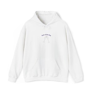 Könnte beinhalten: Weißer Kapuzenpullover mit Fronttasche und Kordelzug. Der Schriftzug "hair of the dog" ist in lila Schrift über einer Grafik von zwei Sektgläsern aufgedruckt. Ein lässiges, bequemes Kleidungsstück.