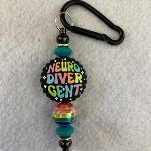 May include: A beaded lanyard with a black carabiner clip. The lanyard features a black, scalloped charm with the words "NEURO DIVERGENT" in colorful letters. Below the charm are rainbow and teal beads, and a small silver keyring.