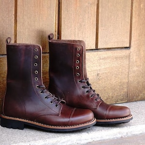 May include: A pair of dark brown leather lace-up boots featuring a cap toe design, a mid-calf height, and a black rubber lug sole. The boots have metal eyelets for the laces and a pull tab at the top of the heel collar for ease of access.