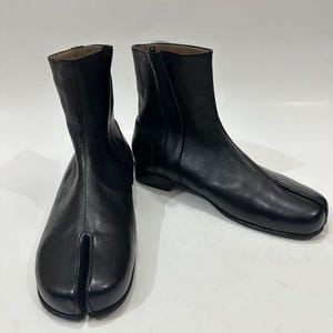 May include: A pair of black leather ankle boots featuring a distinct split-toe design. The boots have a smooth finish, a mid-height shaft, and a side zipper closure. The construction includes a low heel and a clean, minimalist silhouette.