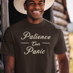 May include: A brown t-shirt with the words "Patience Over Panic" in a cursive font. The person in the image is wearing a white cowboy hat and smiling. The background includes a wooden structure.