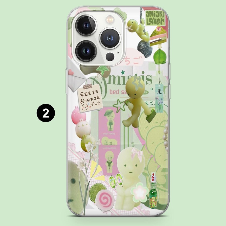 May include: Clear phone case with a whimsical design featuring various green and yellow cartoon figures, a slice of cake, and Japanese text. The case has a transparent border and a light green background.