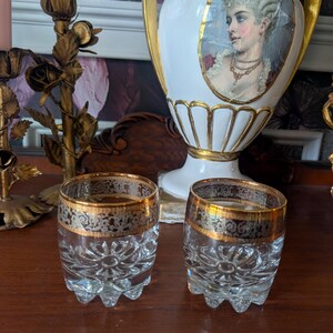 May include: Two clear glass tumblers featuring a cut glass starburst pattern on the base and a wide band of ornate gold and black filigree around the rim. The glasses have a thick, heavy base with a scalloped edge design at the bottom.