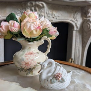 May include: A cream-colored ceramic pitcher featuring a floral pattern filled with artificial pink and cream peonies, accompanied by a white ceramic swan figurine adorned with a small pink rose and green leaves on its side with gold trim accents.