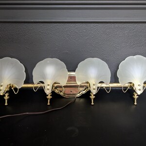 May include: A four-light vanity fixture featuring a horizontal brass bar and four frosted glass shades shaped like scalloped shells. The fixture includes decorative brass hardware and a brown electrical cord extending from the central mounting plate.