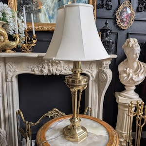 May include: A table lamp with a polished brass base and a flared off-white fabric shade. The lamp features a slender, multi-tiered metal stem with decorative accents and a hexagonal base, providing a steady foundation for the light fixture.