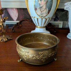 May include: A round, metallic gold-toned bowl featuring an embossed relief pattern of grape clusters and leaves along the exterior rim. The bowl is supported by small, decorative feet and has a smooth, circular interior base with a weathered finish.