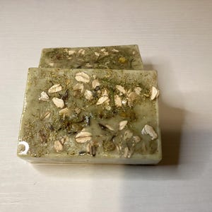 May include: Two rectangular bars of handmade soap. The soap has a light green hue and is embedded with dried herbs and oat flakes. The bars are stacked on top of each other, with the top bar slightly offset.