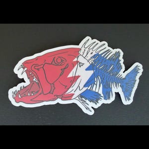 May include: A sticker featuring a stylized fish head with an open mouth and sharp teeth, colored in red, white, and blue. The design transitions from a solid red head to a blue skeletal fish body, incorporating a white lightning bolt shape.