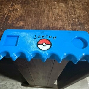 May include: A bright blue, 3D-printed organizer with a series of curved slots along the bottom edge. It features a square and circular indentation, the name "Jayrod", and a Pokeball design.