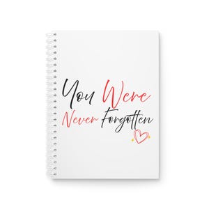 May include: White spiral-bound notebook with the text "You Were Never Forgotten" in black and red script. A small, outlined heart with a gold star is below the text. The notebook has a clean, minimalist design.