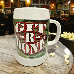 May include: A white ceramic beer mug featuring a green, brown, and black camouflage pattern. The text GIT-R-DONE is printed in large white letters with a red outline. The base of the mug includes the text OFFICIALLY LICENSED BY Larry The Cable Guy.