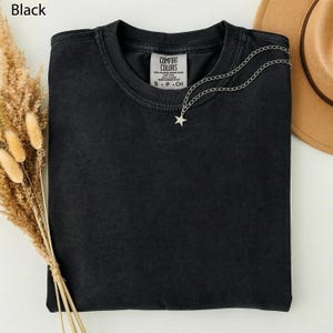 May include: A black Comfort Colors t-shirt with a silver star necklace. The shirt has a small tag with the brand name and size. A brown hat is visible in the background. Dried wheat stalks are on the left side of the image.