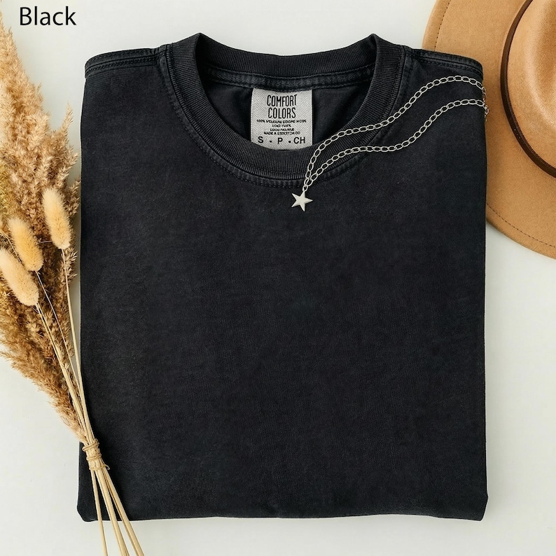 May include: A black Comfort Colors t-shirt with a silver star necklace. The shirt has a small tag with the brand name and size. A brown hat is visible in the background. Dried wheat stalks are on the left side of the image.