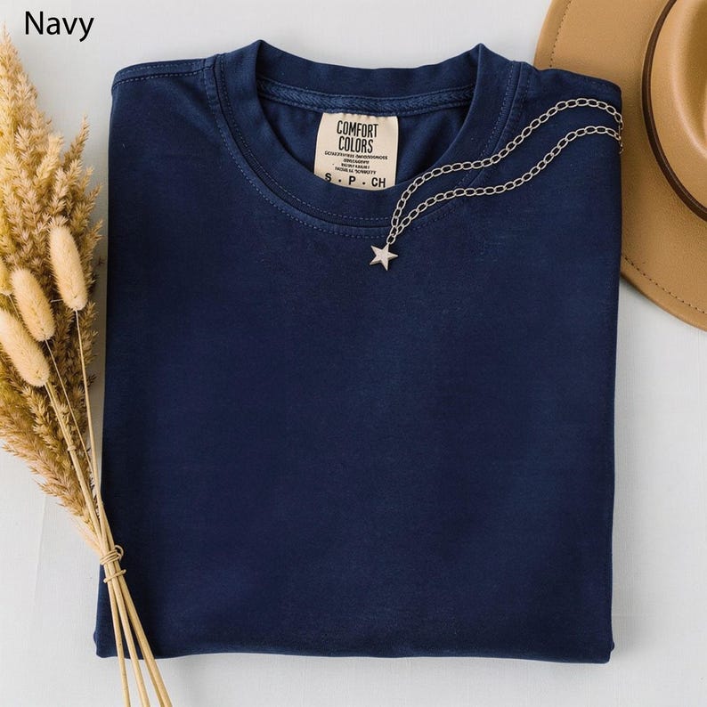 May include: Navy blue t-shirt with a silver star pendant necklace. The shirt has a Comfort Colors tag and is folded neatly. A tan hat is visible in the background. Dried wheat stalks are on the left side of the image.