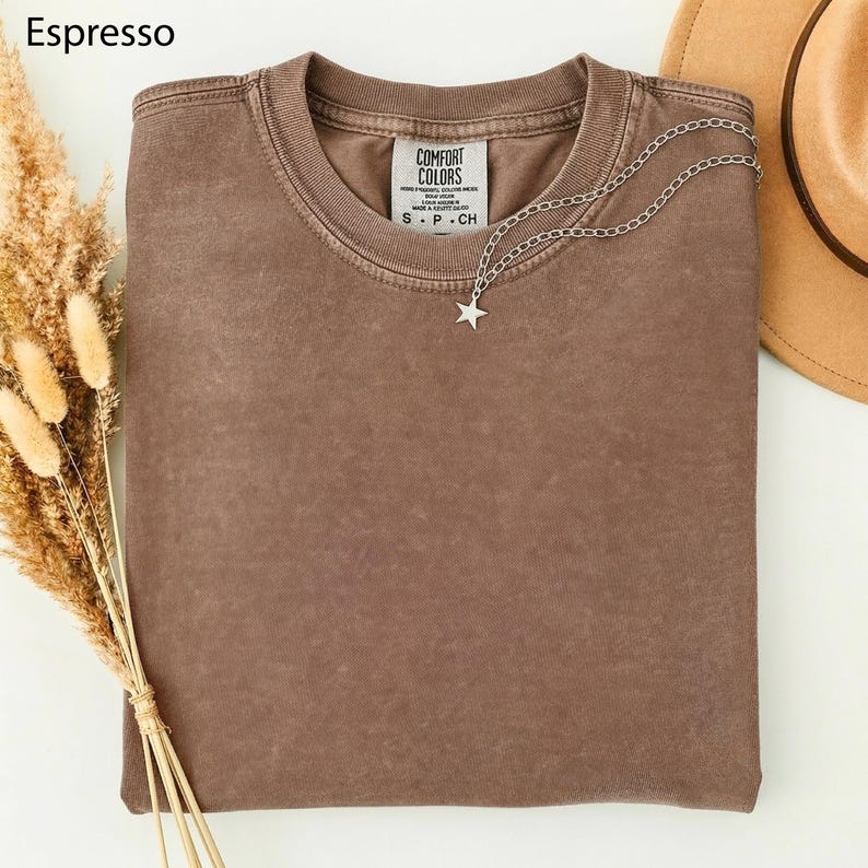 May include: A folded espresso-colored t-shirt with a silver star pendant necklace. The shirt has a Comfort Colors tag. A tan hat and dried wheat are in the background.