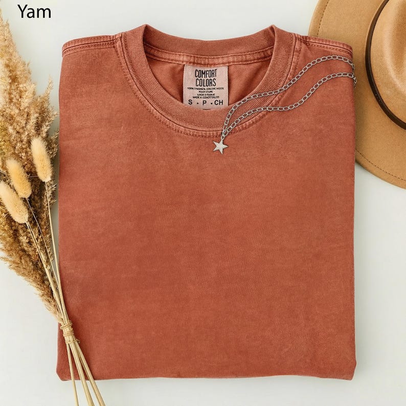 May include: A folded, terracotta-colored t-shirt with a Comfort Colors tag. A silver necklace with a star pendant rests on the shirt. A tan hat is partially visible in the background. Dried wheat stalks are on the left.