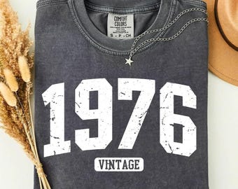 May include: Pepper-colored t-shirt with the year "1976" in large white distressed lettering. The word "VINTAGE" is below the numbers. A silver star pendant necklace and a tan hat are also visible.