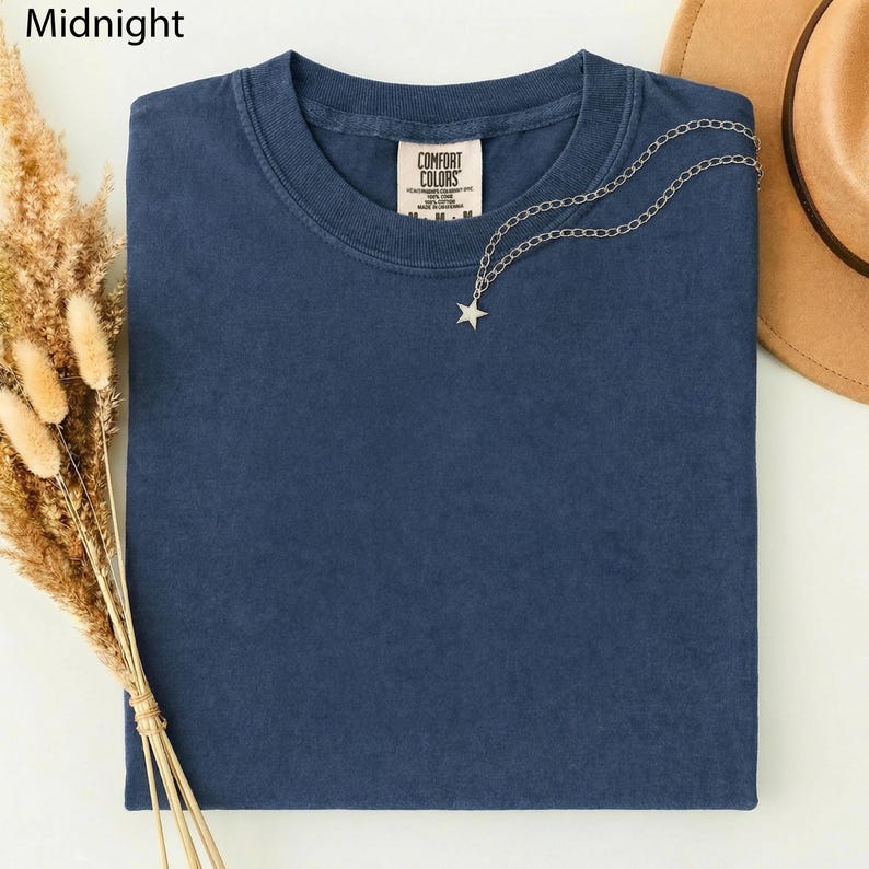 May include: A folded midnight blue t-shirt with a Comfort Colors tag. A silver necklace with a star pendant is draped across the shirt. A tan hat and dried wheat stalks are in the background.