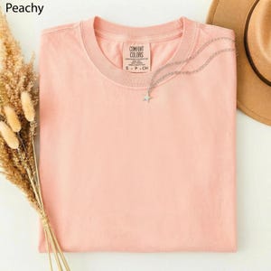 May include: A peach-colored Comfort Colors t-shirt with a silver star pendant necklace. The shirt is folded neatly, with the brand label visible. A brown hat and dried flowers are in the background. The word "Peachy" is in the top left corner.