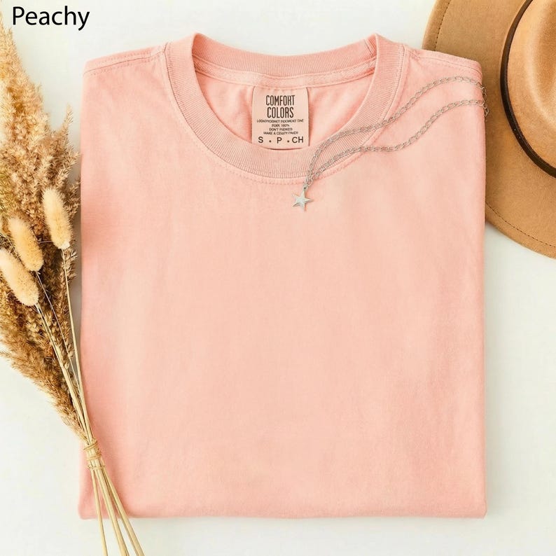 May include: A peach-colored Comfort Colors t-shirt with a silver star pendant necklace. The shirt is folded neatly, with the brand label visible. A brown hat and dried flowers are in the background. The word "Peachy" is in the top left corner.