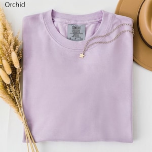 May include: A folded orchid-colored t-shirt with a Comfort Colors tag. A gold star pendant necklace rests on the shirt. A tan hat and dried wheat are in the background.