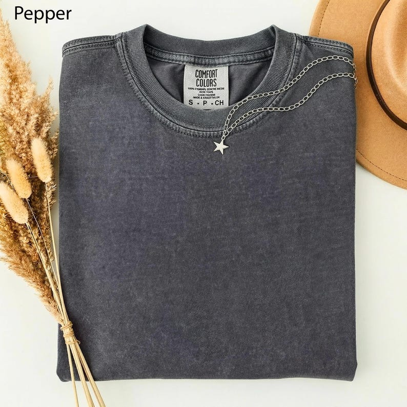 May include: A folded, pepper-colored t-shirt with a silver star pendant necklace. The shirt has a Comfort Colors tag and is next to a brown hat and dried wheat. The shirt is a size S-P-CH.