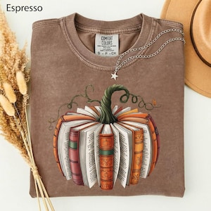 May include: An espresso-colored crew neck t-shirt featuring a graphic of a pumpkin constructed from open books with a green stem. The shirt has a tag labeled Comfort Colors and a silver-toned star pendant necklace resting on the neckline.
