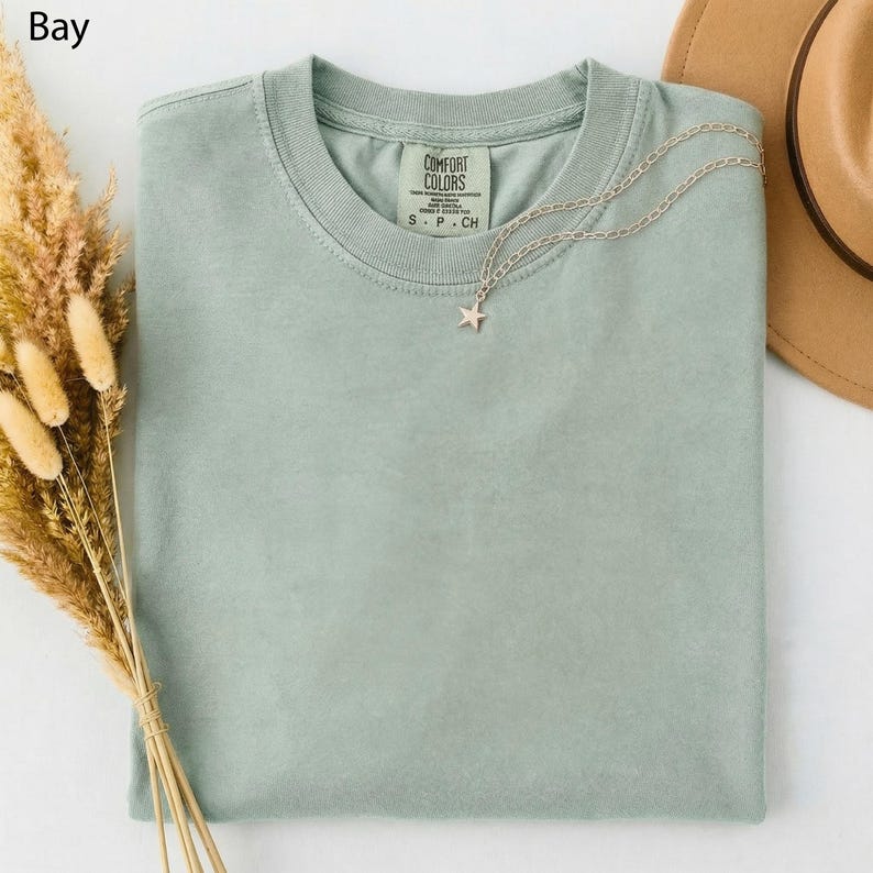 May include: A light green Comfort Colors t-shirt with a star pendant necklace. The shirt has a crew neck and a tag that reads "Comfort Colors". A brown hat and dried flowers are in the background.